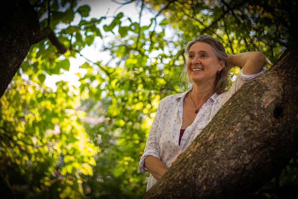 Barbara Rainer, die Gartenarchitektin ist hier in einem Garten mit gutem Gartendesign zu sehen. Sie steht unter einem großen Baum und lächelt dich an.